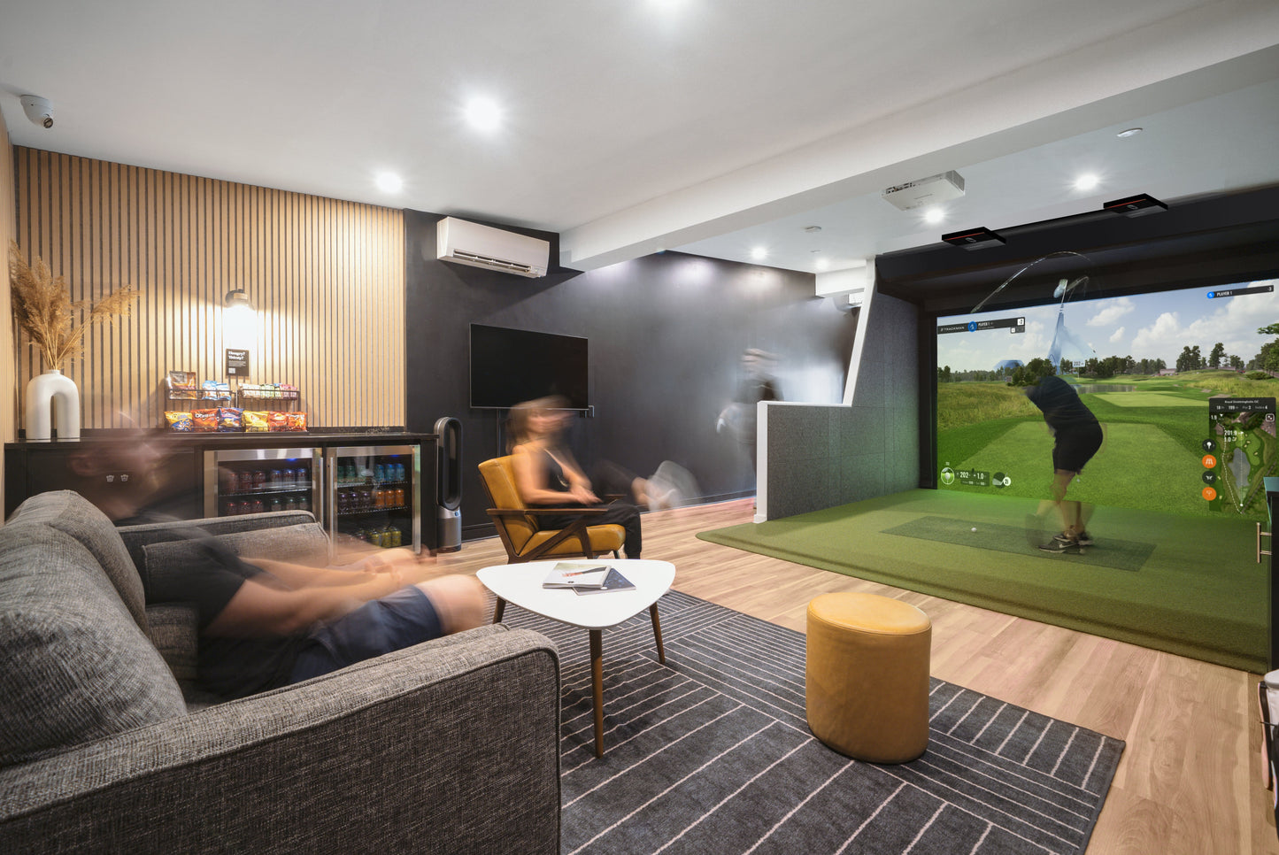 An indoor golf simulator setup with a large screen showing a golf course, a person is seated in front of the screen holding a golf club, with a modern interior design featuring a gray sofa, wooden flooring, and a white accent chair.