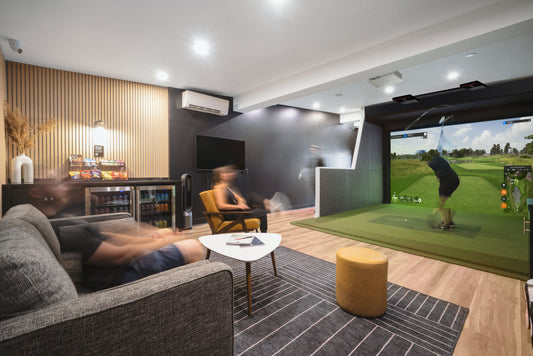 An indoor golf simulator setup with a large screen showing a golf course, a person is seated in front of the screen holding a golf club, with a modern interior design featuring a gray sofa, wooden flooring, and a white accent chair.